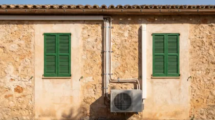 A condenser and its pipe run on the face of a stone farmhouse between two green shuttered windows