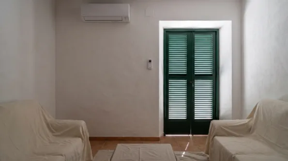 A wall unit above two dust-sheeted armchairs in a closed room, louvred doors shut against the light