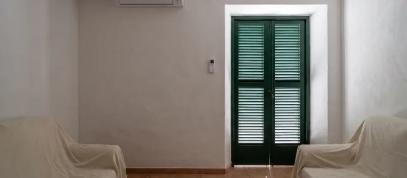 A wall unit above two dust-sheeted armchairs in a closed room, louvred doors shut against the light