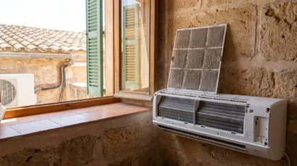 An indoor unit opened at a window, its clogged filter panel lifted clear of the exposed heat exchanger