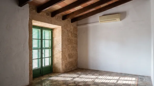 A wall unit under a beamed ceiling in an empty room, light falling through shutters onto tiled floor