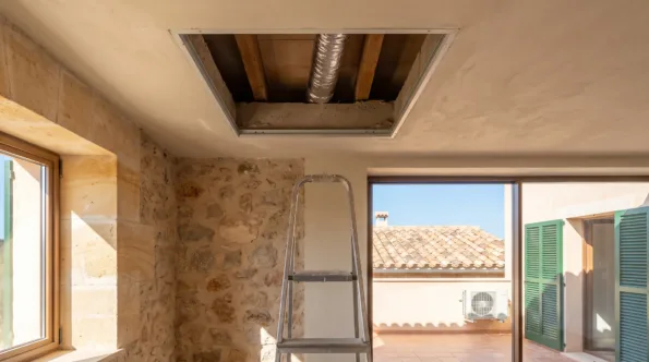 An opened ceiling void with flexible duct above a step ladder, a condenser on the terrace beyond