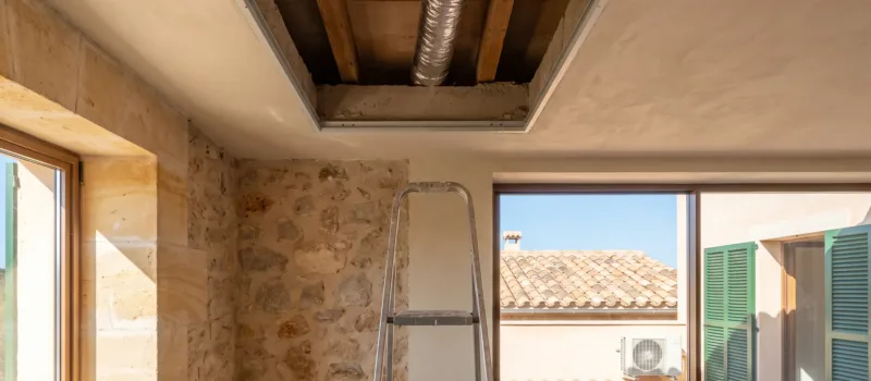 Un falso techo abierto con conducto flexible sobre una escalera de mano, una unidad exterior en la terraza al fondo
