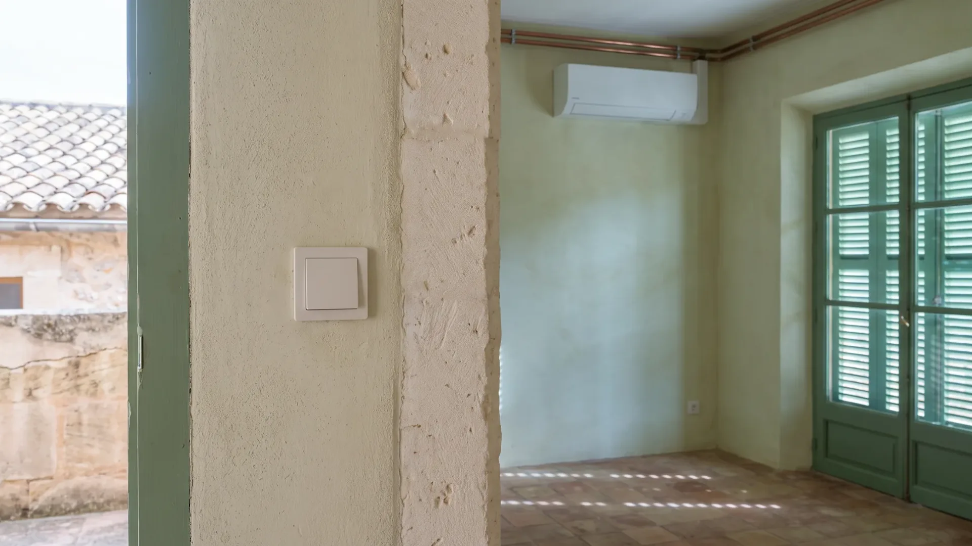 A wall unit and copper pipe run in an empty rendered room, a light switch on a stone reveal nearby
