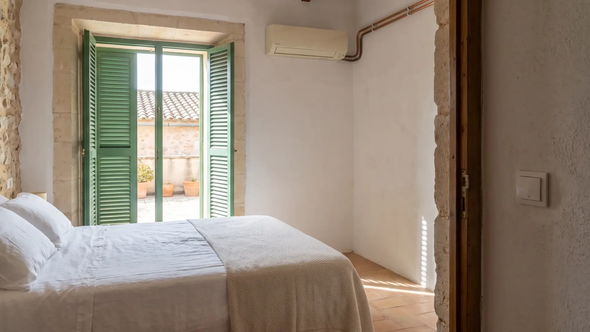 Ein Wandgerät über einem gemachten Bett in einem Raum mit Steinwänden, Läden offen zu einer Terrasse mit Töpfen