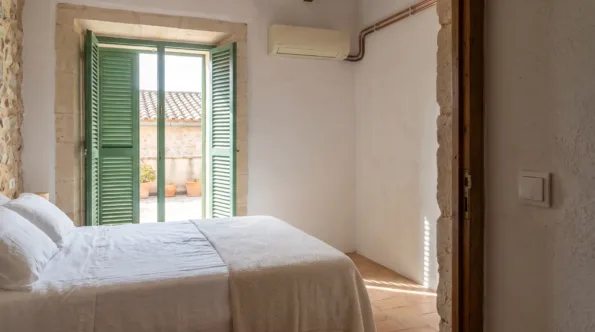 A wall unit above a made bed in a stone walled room, shutters open onto a terrace with pots