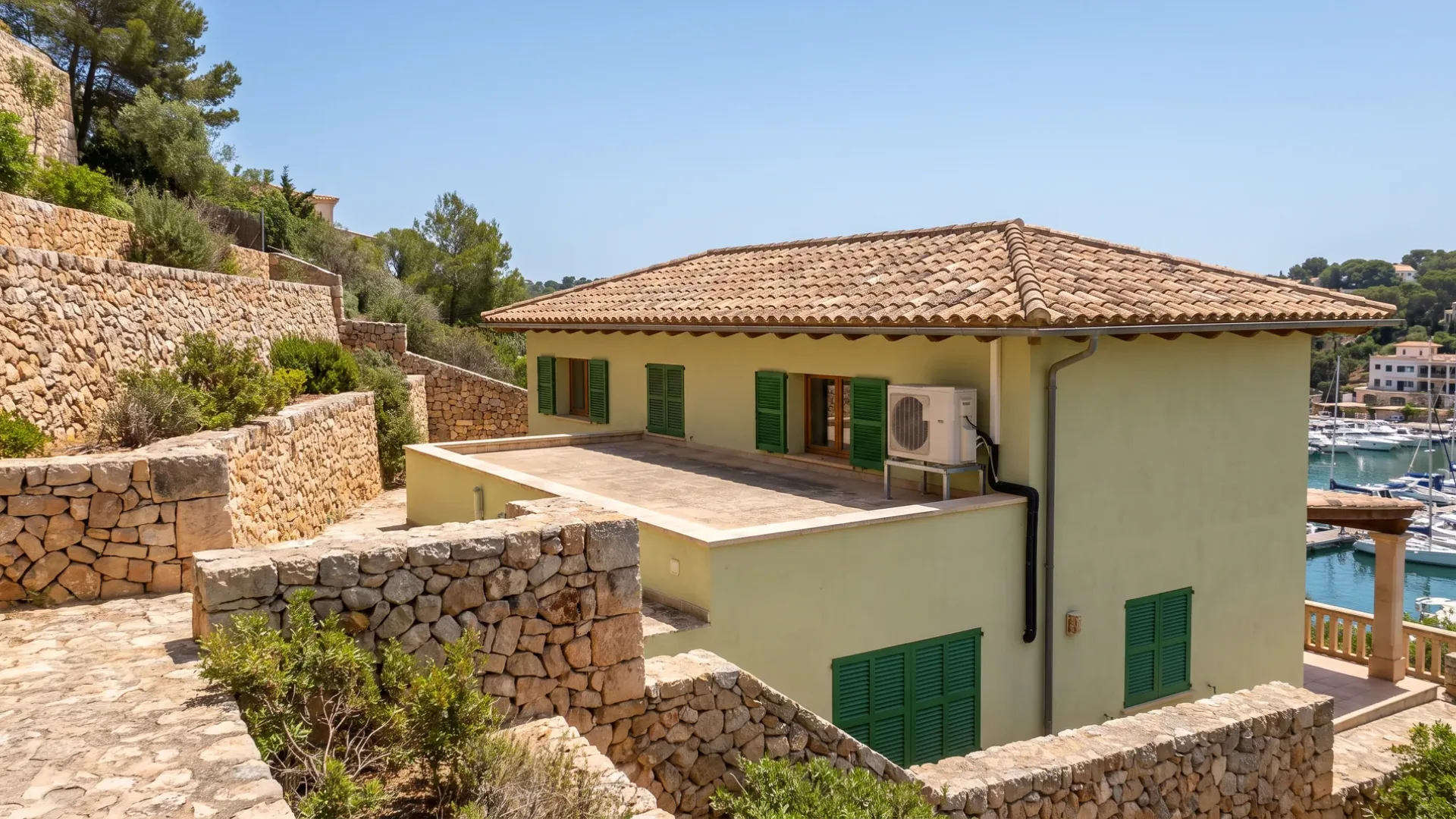 A condenser on the rear wall of a low villa above terraced stone walls, a marina behind it