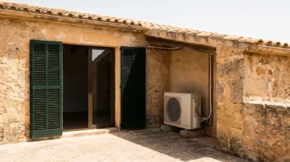 A condenser shaded under a pergola roof beside open shutters and a stone paved terrace