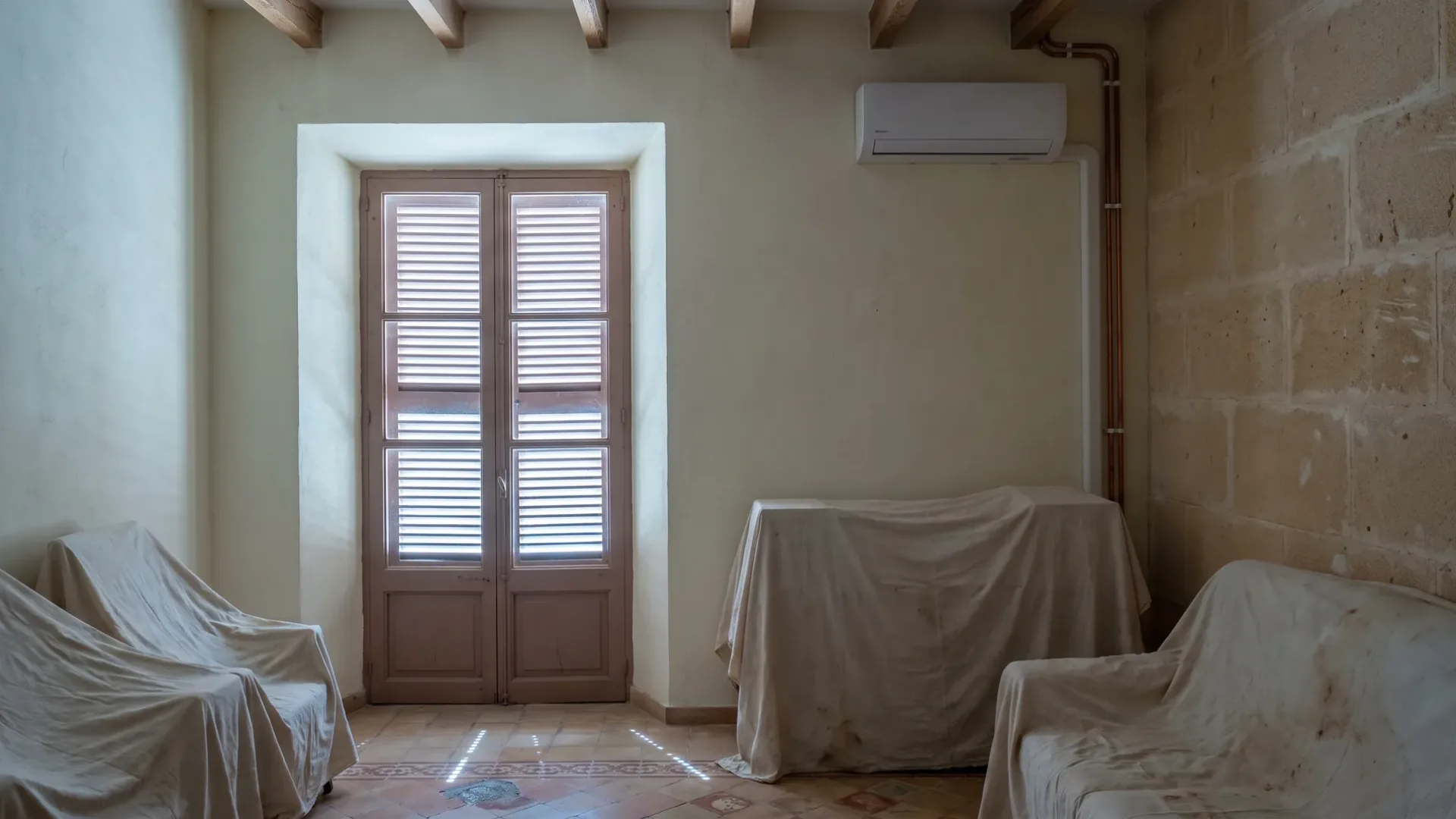 Una unidad de pared sobre muebles cubiertos con sábanas en una habitación con las persianas cerradas, tubería de cobre llevada hasta la esquina