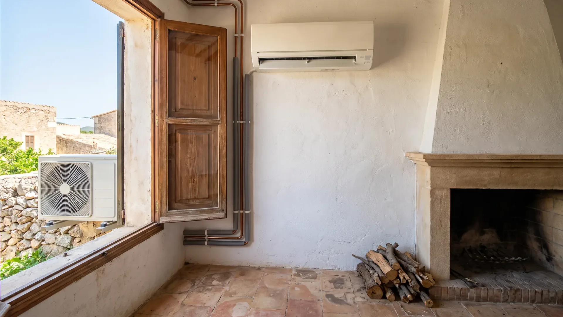A wall unit and its pipe drop beside a stone fireplace, logs stacked and a shuttered window standing open