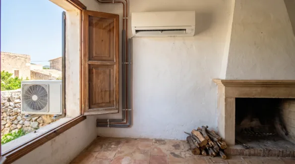 A wall unit and its pipe drop beside a stone fireplace, logs stacked and a shuttered window standing open