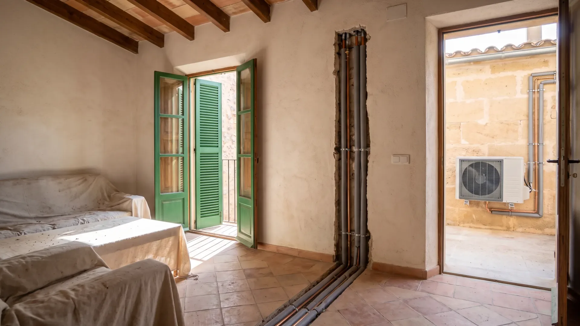 Pipe runs in a chased channel of bare plaster above dust-sheeted furniture, shuttered doors open to a terrace