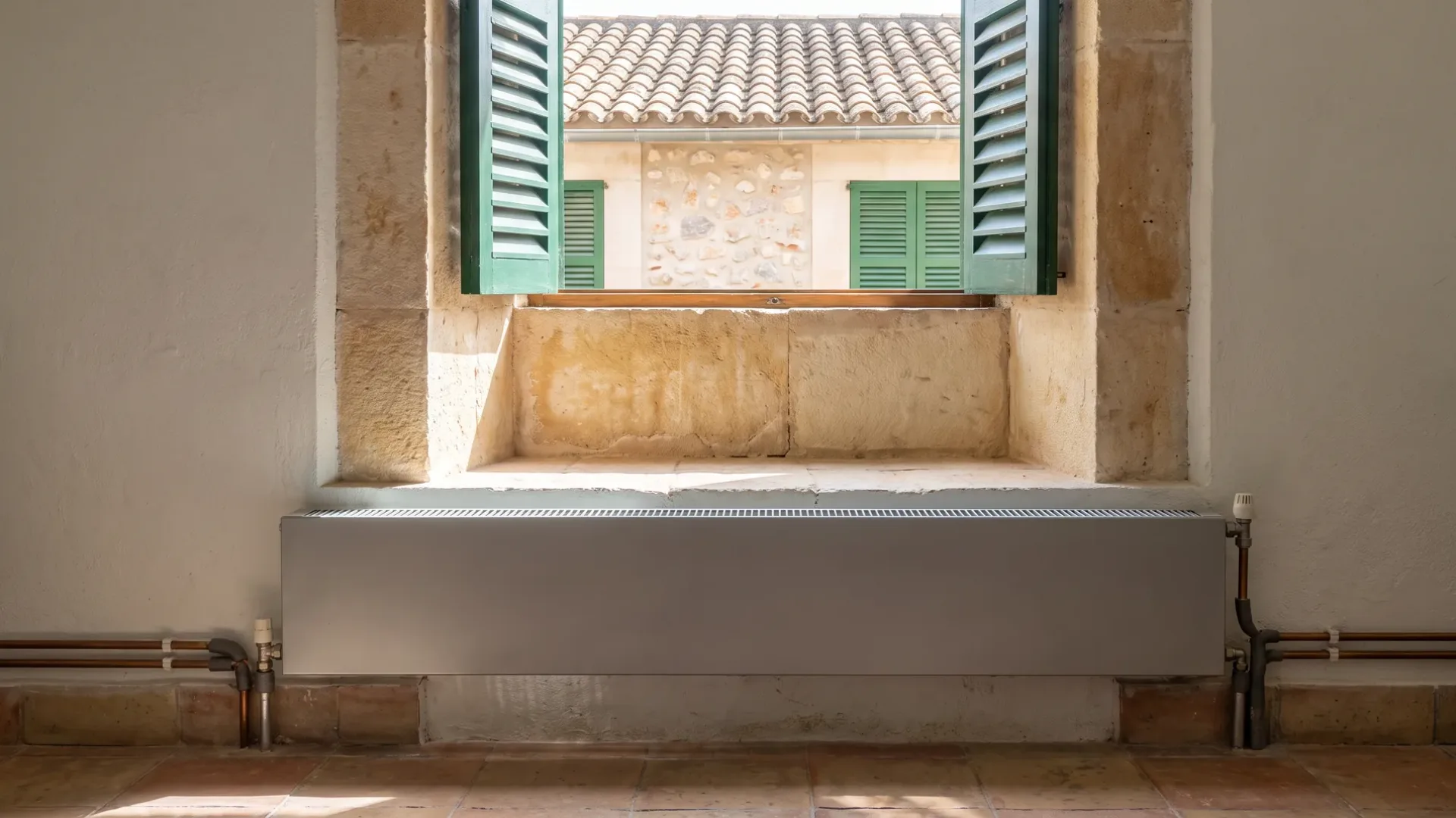 A low panel radiator on copper pipes below a deep stone window recess, green shutters open to a tiled roof