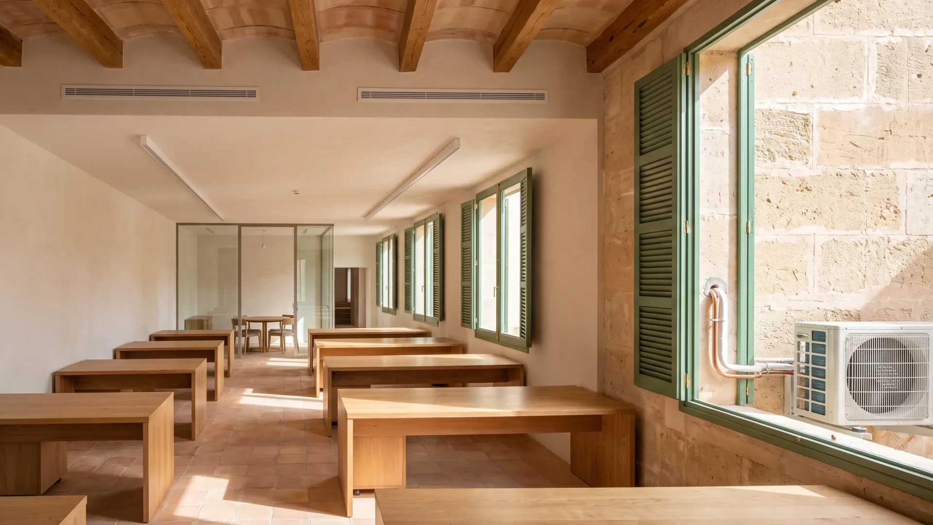 Two linear diffusers in a vaulted beamed ceiling above plain timber desks in a long shuttered workroom